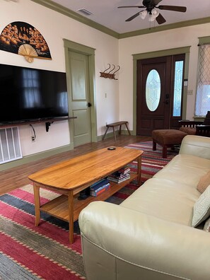 Ein Wohnzimmer mit einem Flachbildfernseher, einem Holztisch, einem beigefarbenen Sofa, einem gemusterten Teppich und einer Tür mit einem Buntglasfenster.