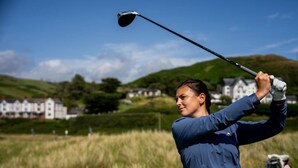 Una persona con chaqueta azul balancea un palo de golf en un campo, con casas al fondo.