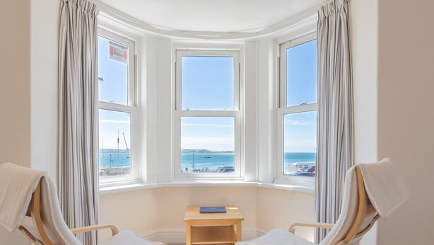 Habitación con tres ventanales que dan a la playa, dos sillas de madera, una mesita de madera y un enchufe en la pared.