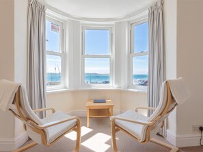 A room with three bay windows overlooking a beach, two wooden chairs, a small wooden table, and a power outlet on the wall. - 1 Bed in Aberdyfi (oc-dy019) (Aberdovey)