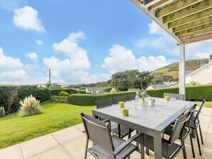 Um pátio externo com mesa e cadeiras, rodeado por um jardim bem cuidado e vista para o campo.