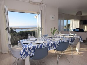 Uma área de jantar com uma mesa posta para quatro pessoas, uma planta em vaso e vista para o mar.