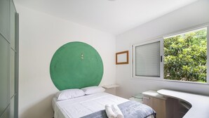 Quarto com cama, cabeceira circular verde, janela com vista para vegetação, mesa de cabeceira e pequena área de trabalho.