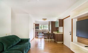 Ein modernes Wohnzimmer mit einem grünen Sofa, einer Küche mit Holzschränken und einem Fernseher an der rechten Wand.