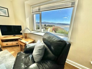 A living room with a black leather sofa, a TV on a wooden stand, a small table with a lamp, and a large window with a view of the sea and hills. - 3 Bed in Aberdyfi (oc-dy024) (Aberdovey)