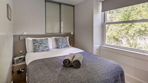 Ein ordentlich bezogenes Bett mit gemusterten Kissen, ein Nachttisch mit Radio und ein Fenster mit Blick ins Grüne.
