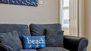 Ein Zimmer mit einem grauen Sofa, einem blauen Kissen mit Strand- und Sternsymbolen sowie einem Fischgemälde an der Wand.
