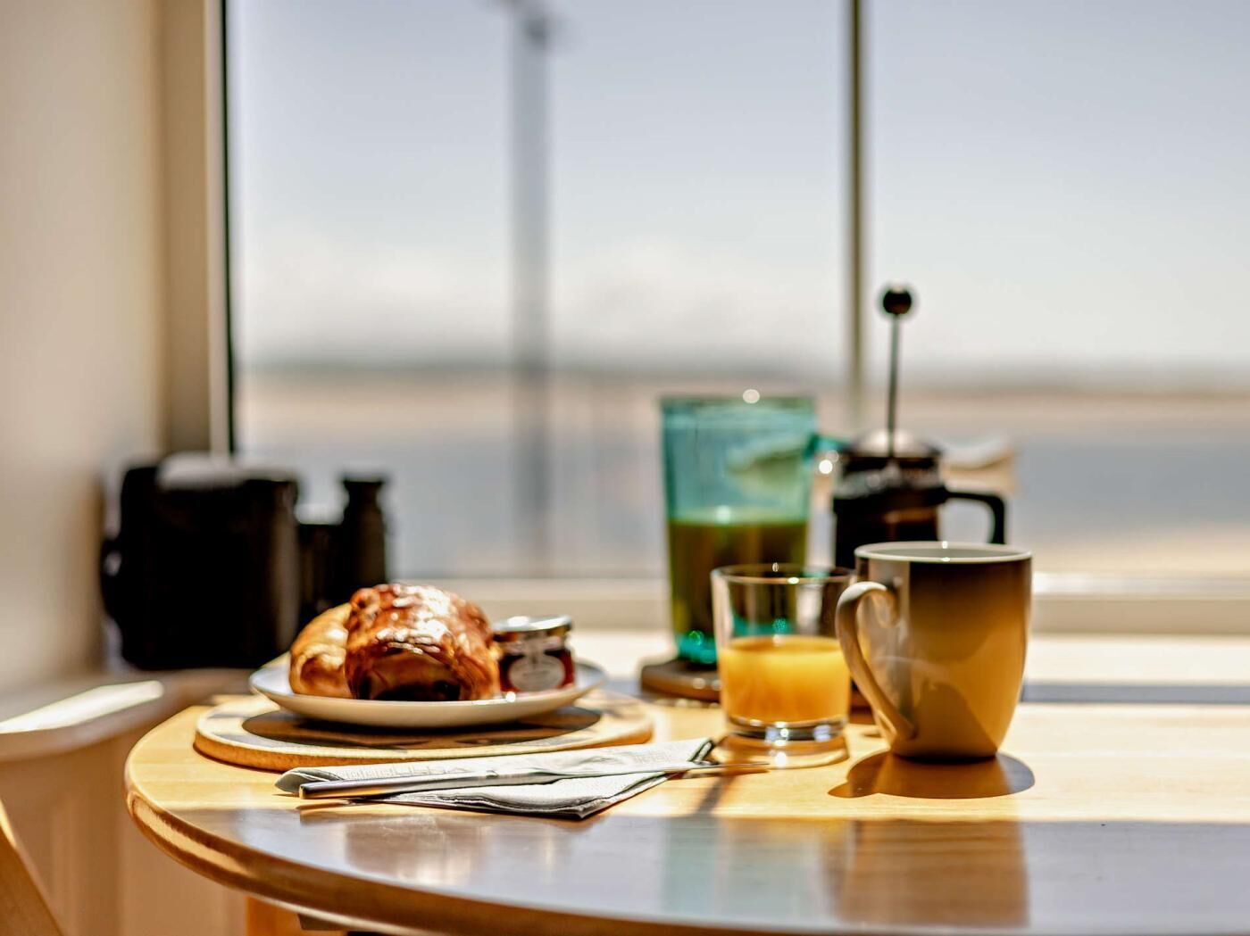 Une table dressée pour le petit-déjeuner, avec un croissant, un verre de jus d’orange et une cafetière, le tout placé près d’une fenêtre.
