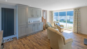 A modern kitchen with grey cabinets and a wooden floor, a seating area with two chairs, and a large window with curtains. - 2 Bed in Aberdyfi (oc-dy049) (Aberdovey)