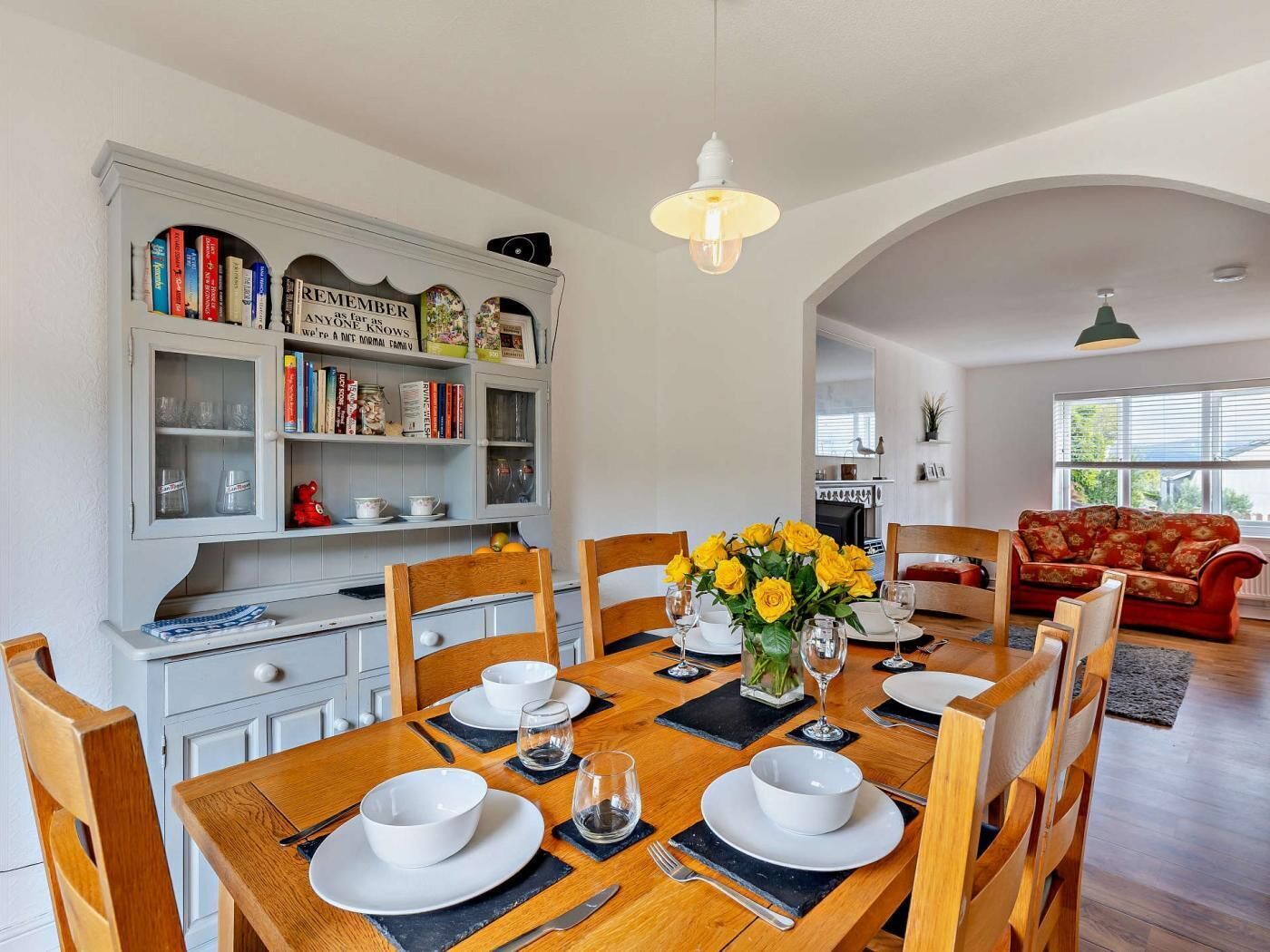 Un coin repas avec une table en bois, dressée pour quatre personnes, entourée de chaises. Une bibliothèque contenant divers objets se trouve à gauche.