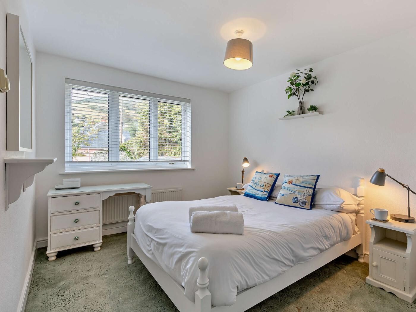 A bedroom with a bed, bedside tables, a desk, a window with blinds, and a wall-mounted shelf with a plant.
