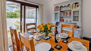 A dining area with a table set for four, a bookshelf with books, and a view through a glass door. - 3 Bed in Aberdyfi (oc-dy048) (Aberdovey)