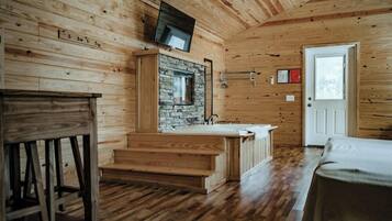 A wooden cabin interior with a stone fireplace, a wooden staircase, a sink, and a television mounted on the wall.