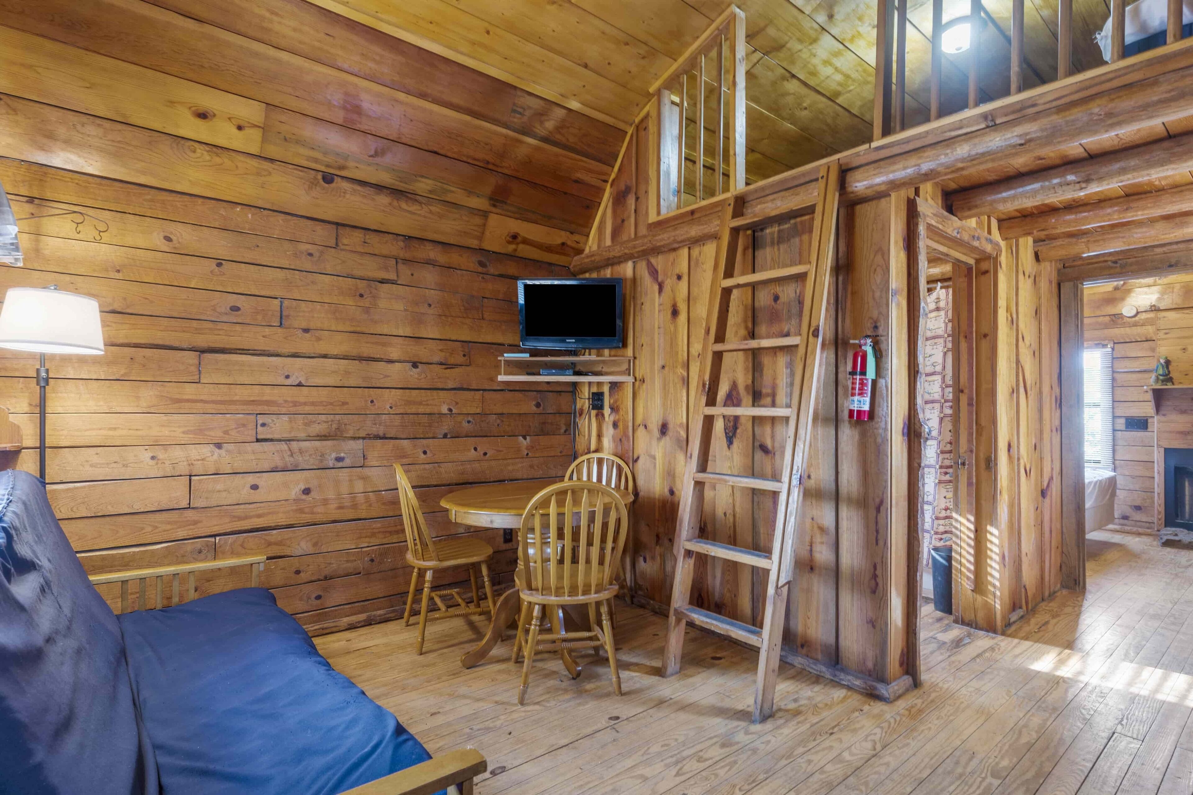 A wooden cabin interior with a TV, a dining table, chairs, a blue sofa, and a wooden ladder.
