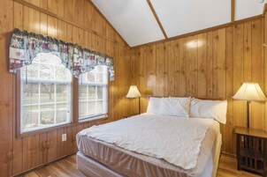 Una habitación con paneles de madera, una cama, dos ventanas con persianas y dos lámparas de noche.