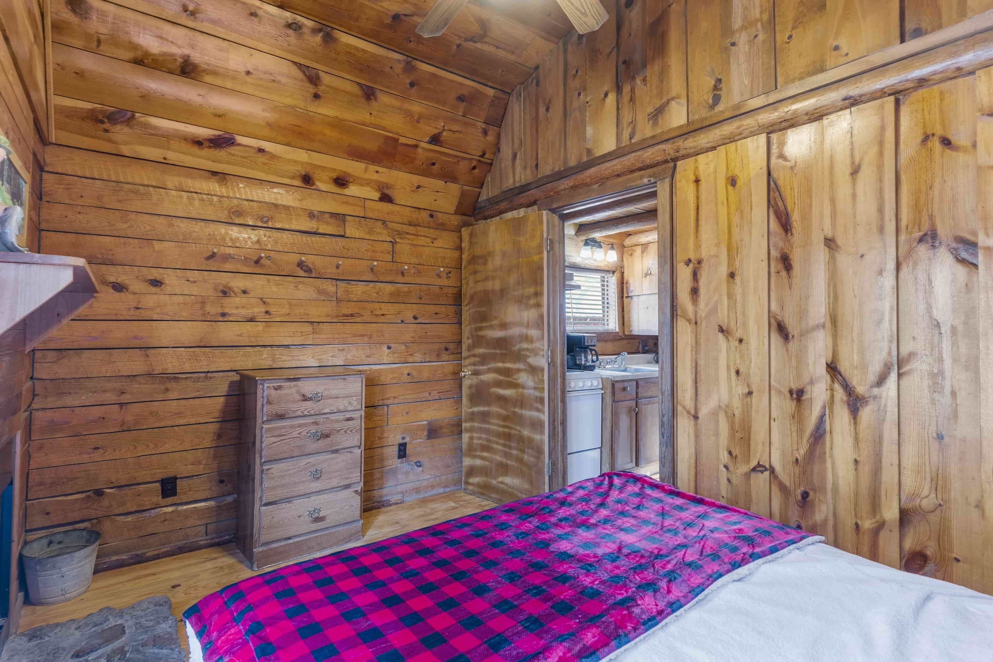 Une pièce de style cabane en bois, comprenant un lit, une commode en bois, un petit coin cuisine et une porte donnant sur une autre pièce.