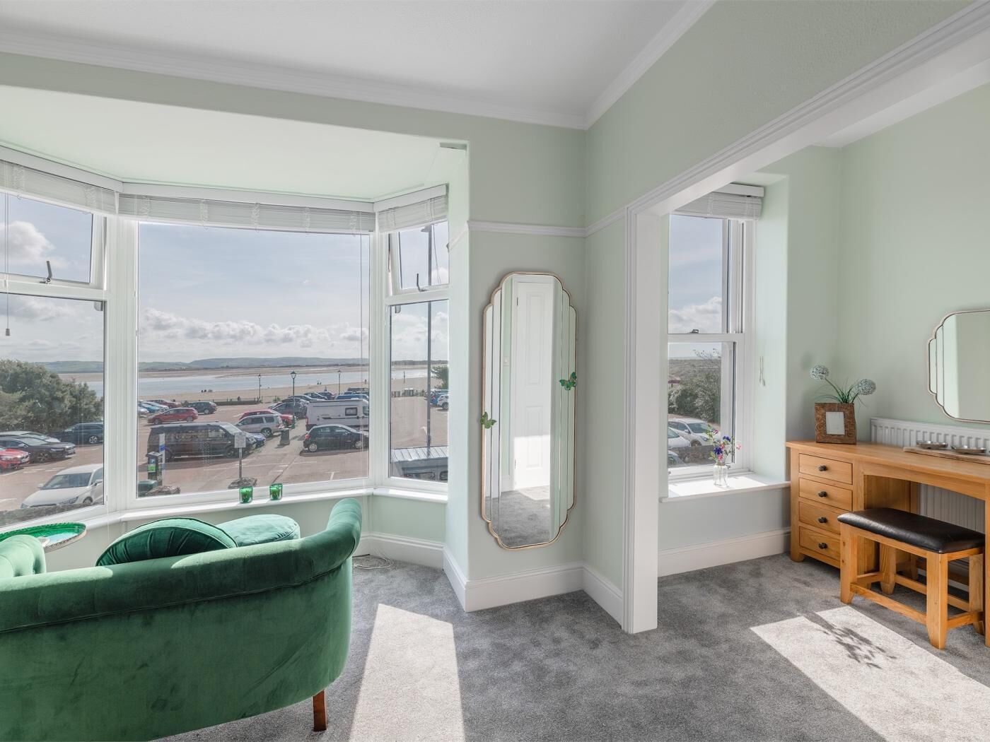 A room with a bay window, a green armchair, a wooden dresser, and a mirror.