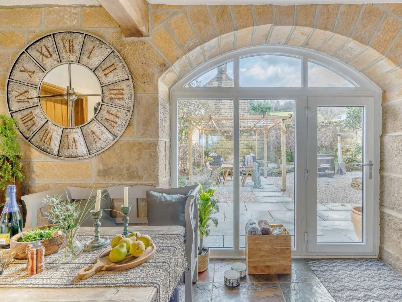 A room with a stone wall, a large clock, a table with fruits and candles, and a view of an outdoor patio.