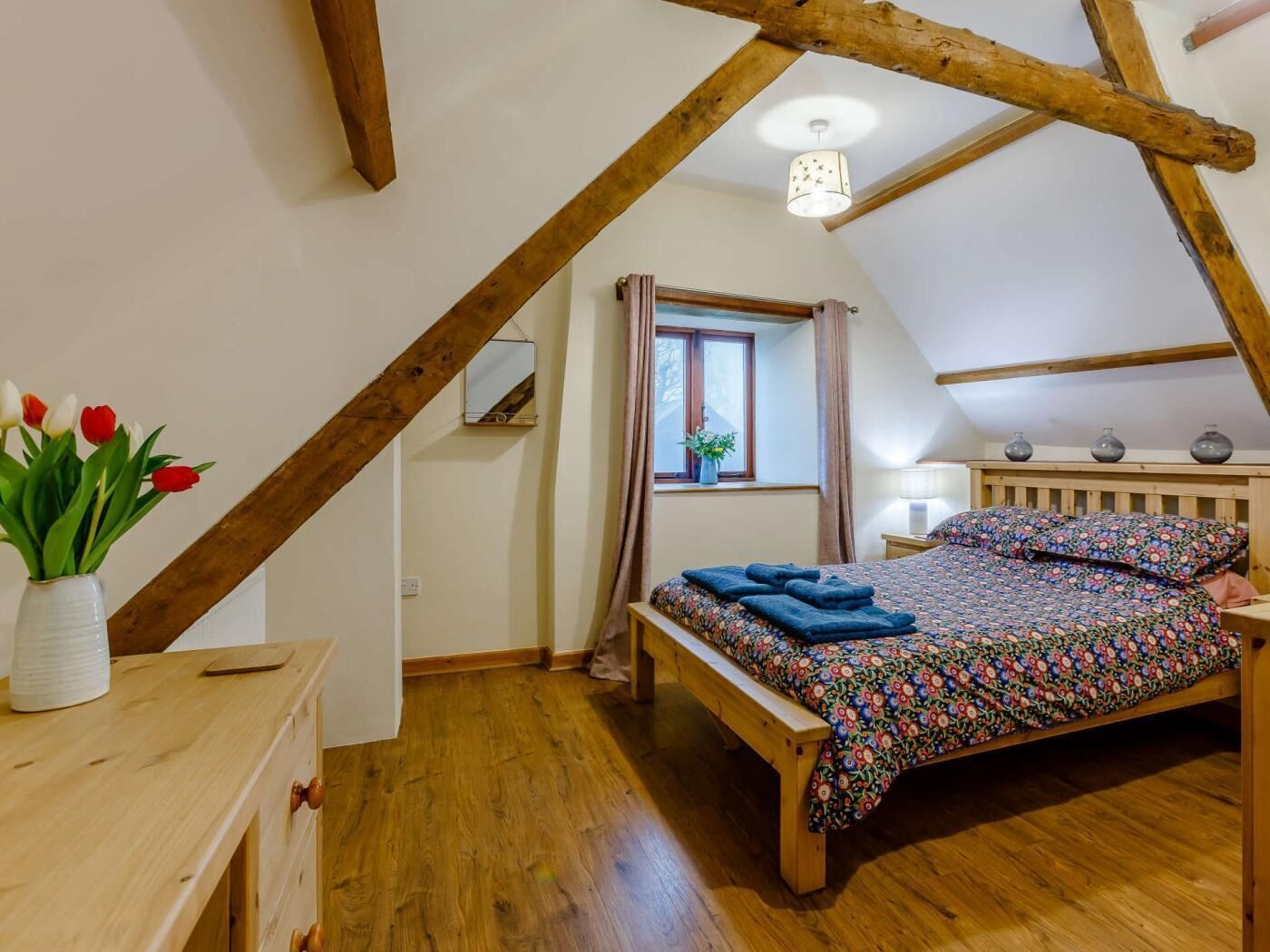 Ein Schlafzimmer mit einem hölzernen Bett, einem Nachttisch, einem Fenster mit Vorhängen, einer Holzbalkendecke und einer Vase mit Blumen.