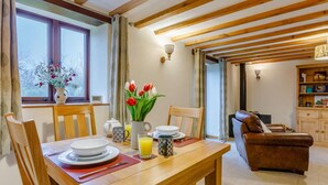 A dining area with a wooden table, chairs, and a vase of flowers. There is a window with curtains, a bookshelf, and a leather sofa in the background. - 1 Bed in Bideford (oc-86571) (Hartland)
