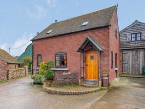 A red brick house with a wooden door, a small porch with a table and chairs, and a green fence in the background.