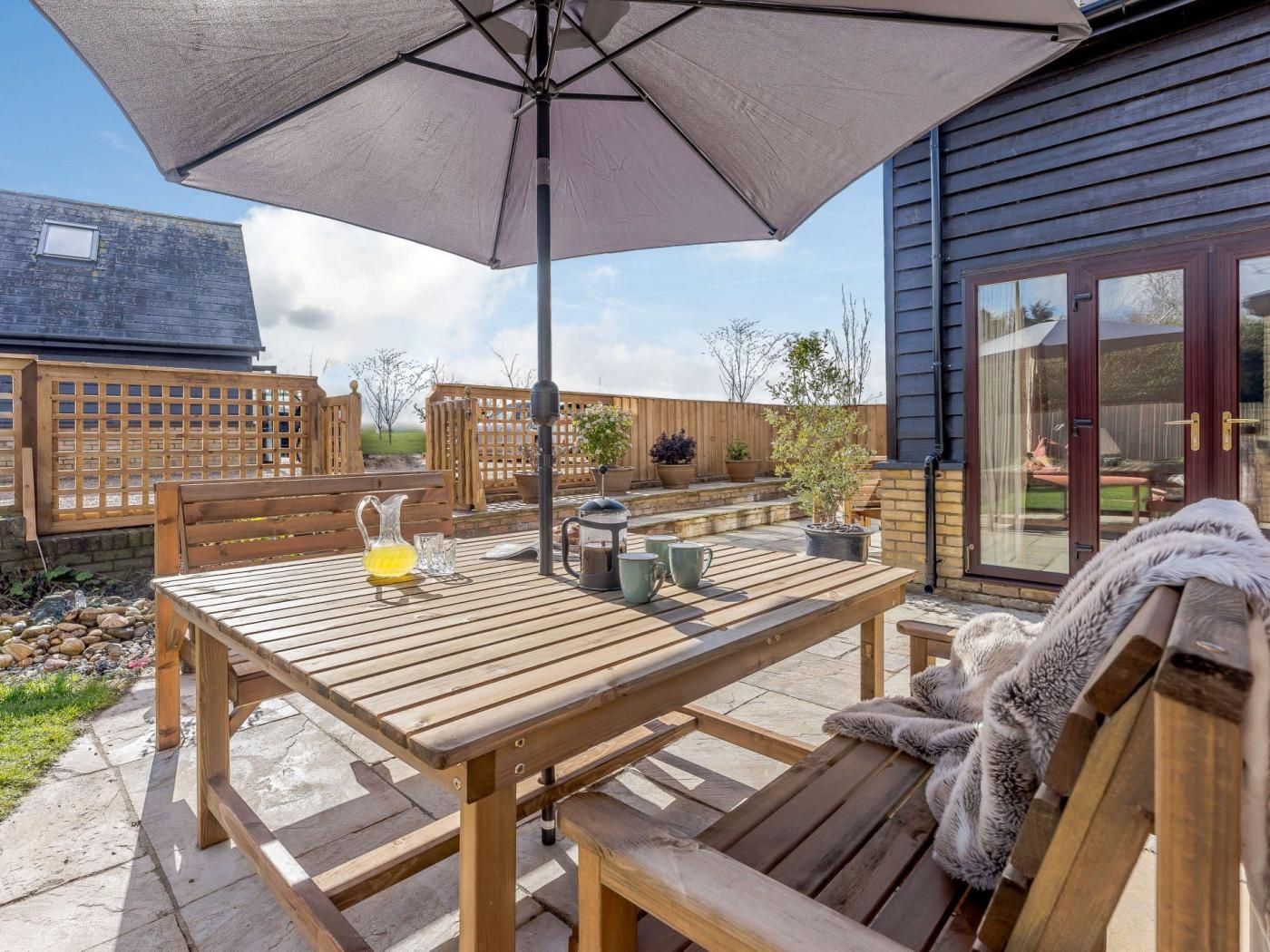 Une terrasse extérieure avec une table et des bancs en bois, un parasol et une vue sur une maison à l’extérieur sombre et dotée de portes vitrées.