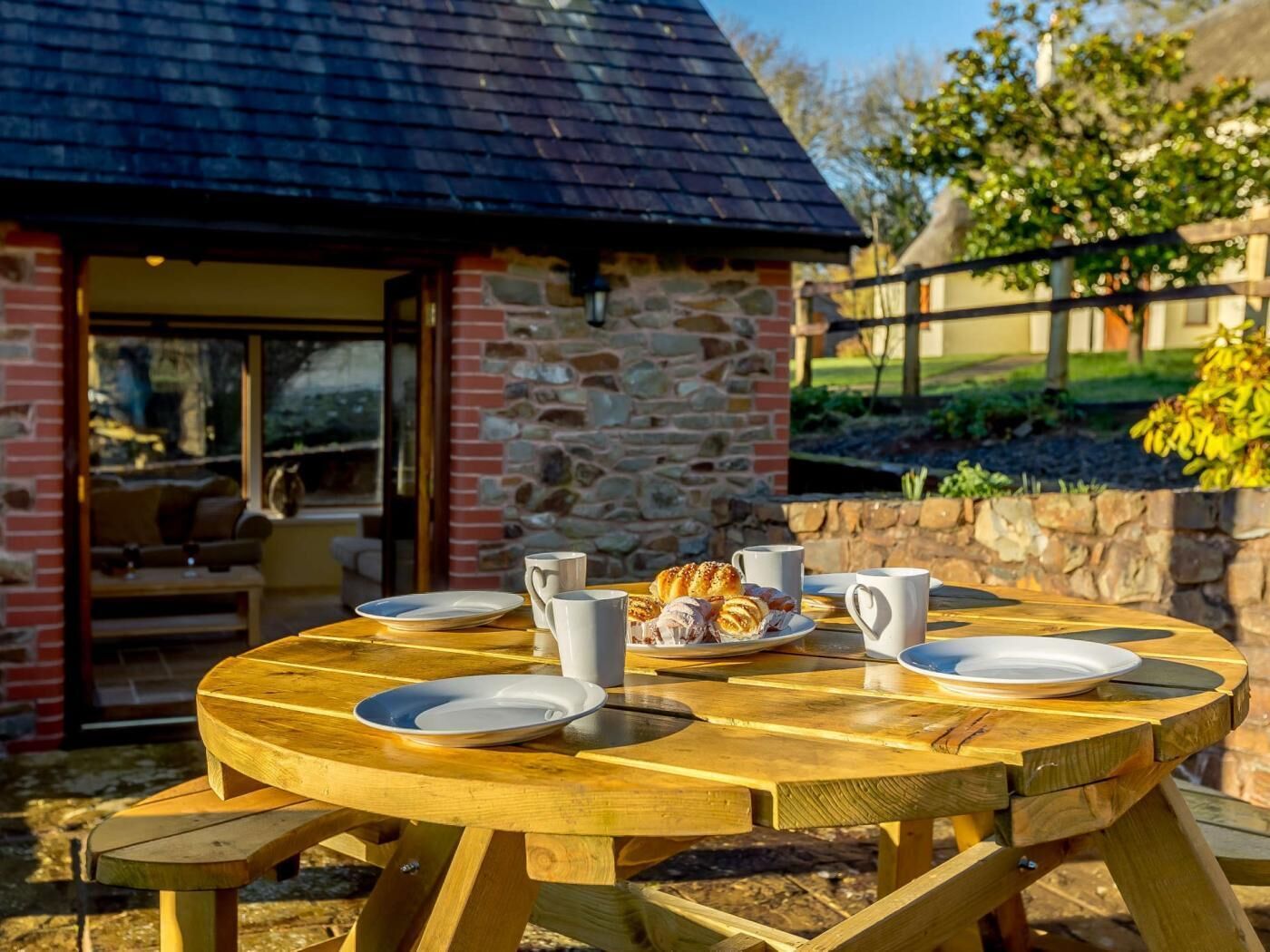 A wooden picnic table with plates, cups, and pastries, set outdoors with a stone building in the background.