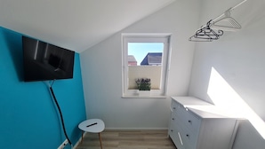 Ein Zimmer mit an der Wand montiertem Fernseher, einem kleinen Tisch, einem Fenster mit Jalousien und einer weißen Kommode.