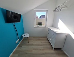 A room with a television mounted on the wall, a small table, a window with blinds, and a white dresser.