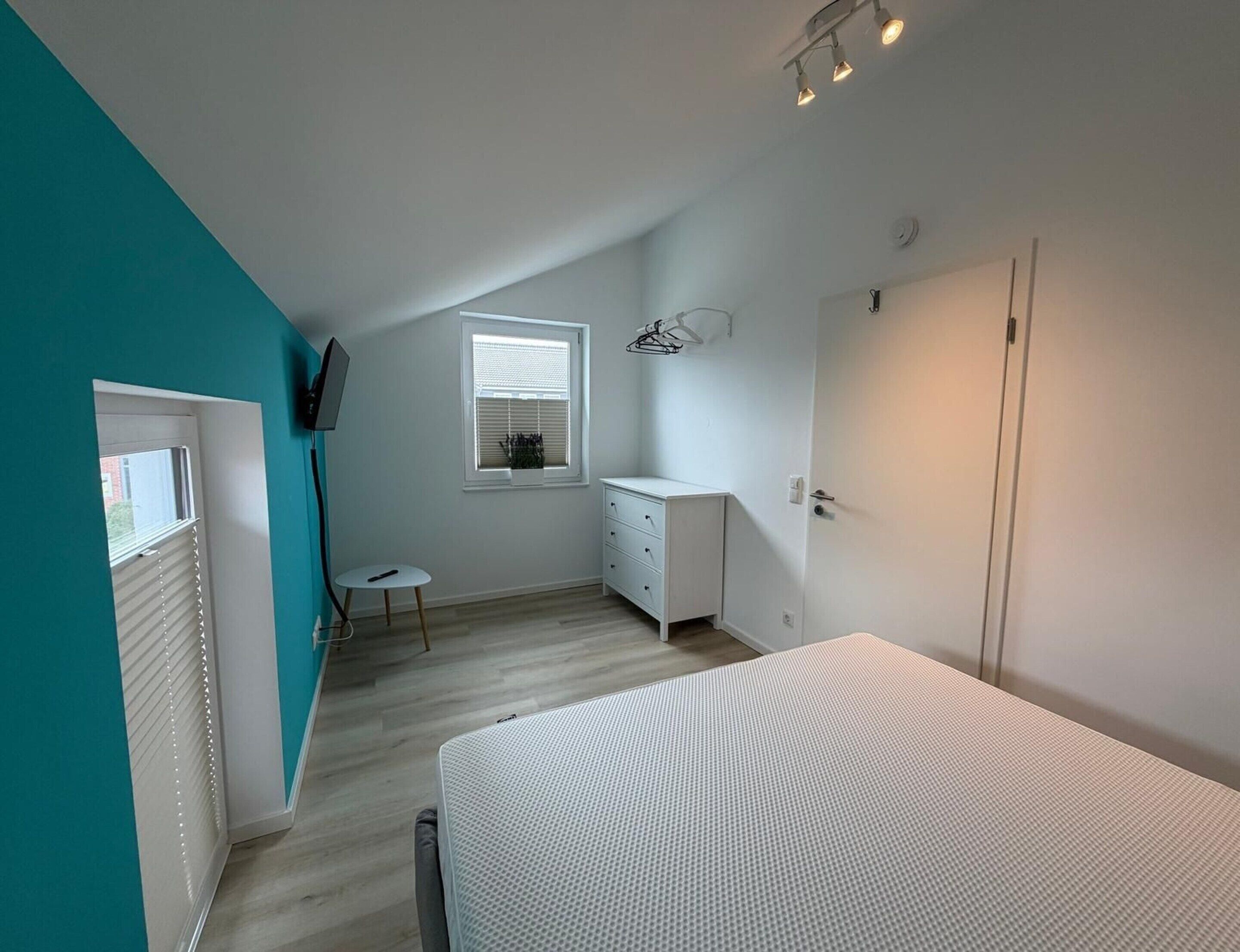 A bedroom with a bed, a dresser, a window with blinds, a small table, and a TV mounted on the wall.