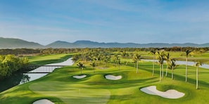 Een golfbaan met zandbunkers, palmbomen en een waterhindernis.