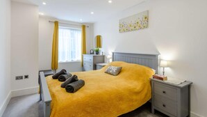 Une chambre à coucher comprenant un lit, des tables de chevet, une commode, une fenêtre avec des rideaux et une œuvre d’art accrochée au mur.