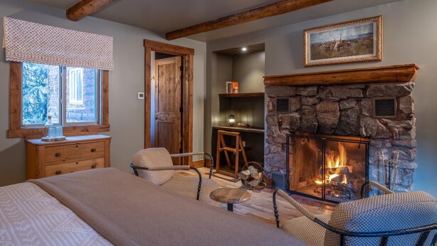 A cozy room with a stone fireplace, wooden beams, a bed, a window with a view, and a bookshelf. - Tumbling River Ranch (Grant)