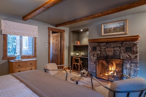A cozy room with a stone fireplace, wooden beams, a bed, a window with a view, and a bookshelf.