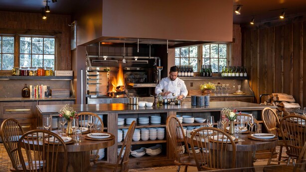 A chef preparing food in a kitchen with a large window, wooden furniture, and a dining area set for guests.