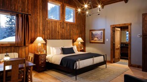 A wooden interior with a bed, two bedside lamps, a dining area with a table and chairs, and a framed picture on the wall.