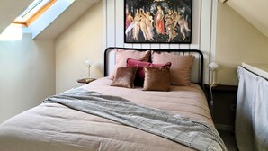 Ein Schlafzimmer mit schräg gedachter Decke, einem Bett mit gestreifter Decke, zwei Nachttischlampen und einem Gemälde über dem Bett.