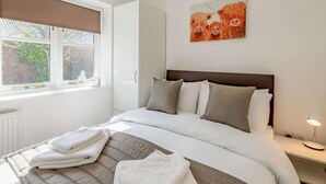 A hotel room with a bed, pillows, a bedside table with a lamp, a window with blinds, and a painting of Highland cattle on the wall. - 2 Bed in Louth (oc-86797) (Fotherby)