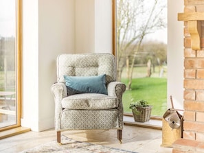 A corner room with a patterned armchair, a blue cushion, a view of a garden, a brick fireplace, and a wicker basket. - 3 Bed in Pershore (oc-86760) (Eckington)