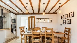 Een eetgedeelte met een houten tafel en stoelen, een keuken met moderne apparatuur, en een plafond met zichtbare balken en hangende lampen.