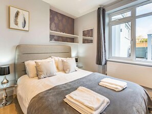 Ein ordentlich bezogenes Bett mit Kissen und Decke, ein Nachttisch mit Lampe und ein Fenster mit Vorhängen.