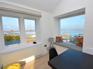 Ein Zimmer mit großem Fenster, das einen Parkplatz und ein Gewässer blickt, mit Tisch und Sitzbank mit Kissen.