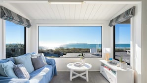 Una habitación con sofá, una mesita, un armario blanco y ventanas amplias con vistas al mar.