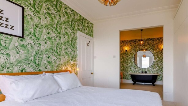 A bedroom with a bed, a door, and a bathroom with a bathtub and a mirror. - 6 Bed in Ilfracombe (oc-86780) (Ilfracombe)