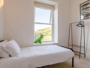 Uma cama de solteiro com roupa de cama branca, uma janela com vista para uma paisagem verde e um suporte de metal preto com uma toalha de mão.