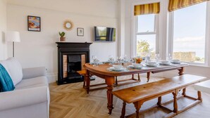 A dining area with a wooden table set for four, a fireplace, a television, and a view of the outdoors. - 6 Bed in Ilfracombe (oc-86780) (Ilfracombe)