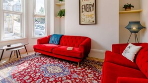 A living room with a red sofa, a patterned rug, a framed motivational poster, and a view of the outside through large windows. - 6 Bed in Ilfracombe (oc-86780) (Ilfracombe)