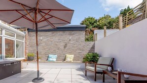 A patio with a tiled floor, a wooden bench, a table with cups, and a large umbrella. - 6 Bed in Ilfracombe (oc-86780) (Ilfracombe)