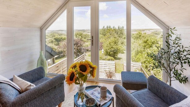 Ein Zimmer mit schrägem Dach, großen Fenstern, zwei grauen Sofas, einem kleinen Tisch mit einer Vase voller Sonnenblumen und Blick auf Grünflächen sowie eine Holzterrasse vor der Tür.