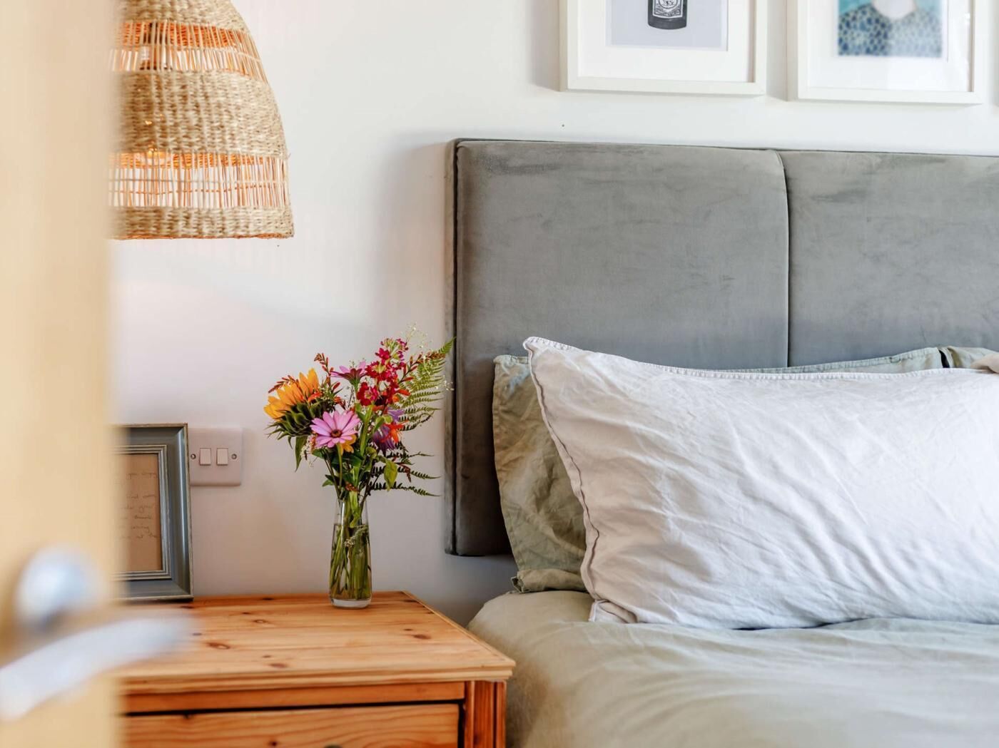 A bedroom with a bed, a bedside table with a lamp, a framed picture, and a vase with flowers.